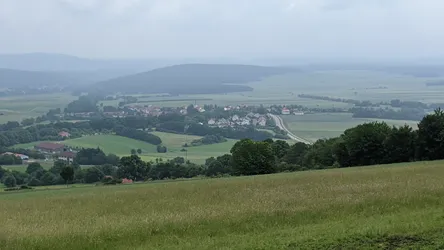 Weiter Blick von der Höhe auf ein Dorf im Tal, dahinter Felder und bewaldete Rücken
