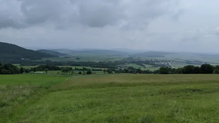 Weite Rhönlandschaft unter Wolken: Wiesen, Waldstücke und ein Ort am Fuß der Höhen