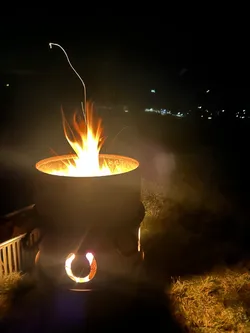 Feuertonne mit ausgeschnittenem Hufeisen brennt in der Nacht, unten im Tal die Lichter der Orte