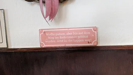 Rotes Blechschild mit einem Spruch übers Putzen, darüber ein Kranz mit rot-weiß kariertem Band