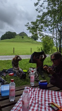 Picknick am Fuß des Wachtküppels: drei aus der Gruppe an Decke und Holztisch, dahinter der Berg mit dem Gipfelkreuz