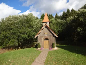 Kleine Holzkapelle mit rotem Spitzturm am Waldrand, davor ein gepflasterter Weg und Blumenkübel