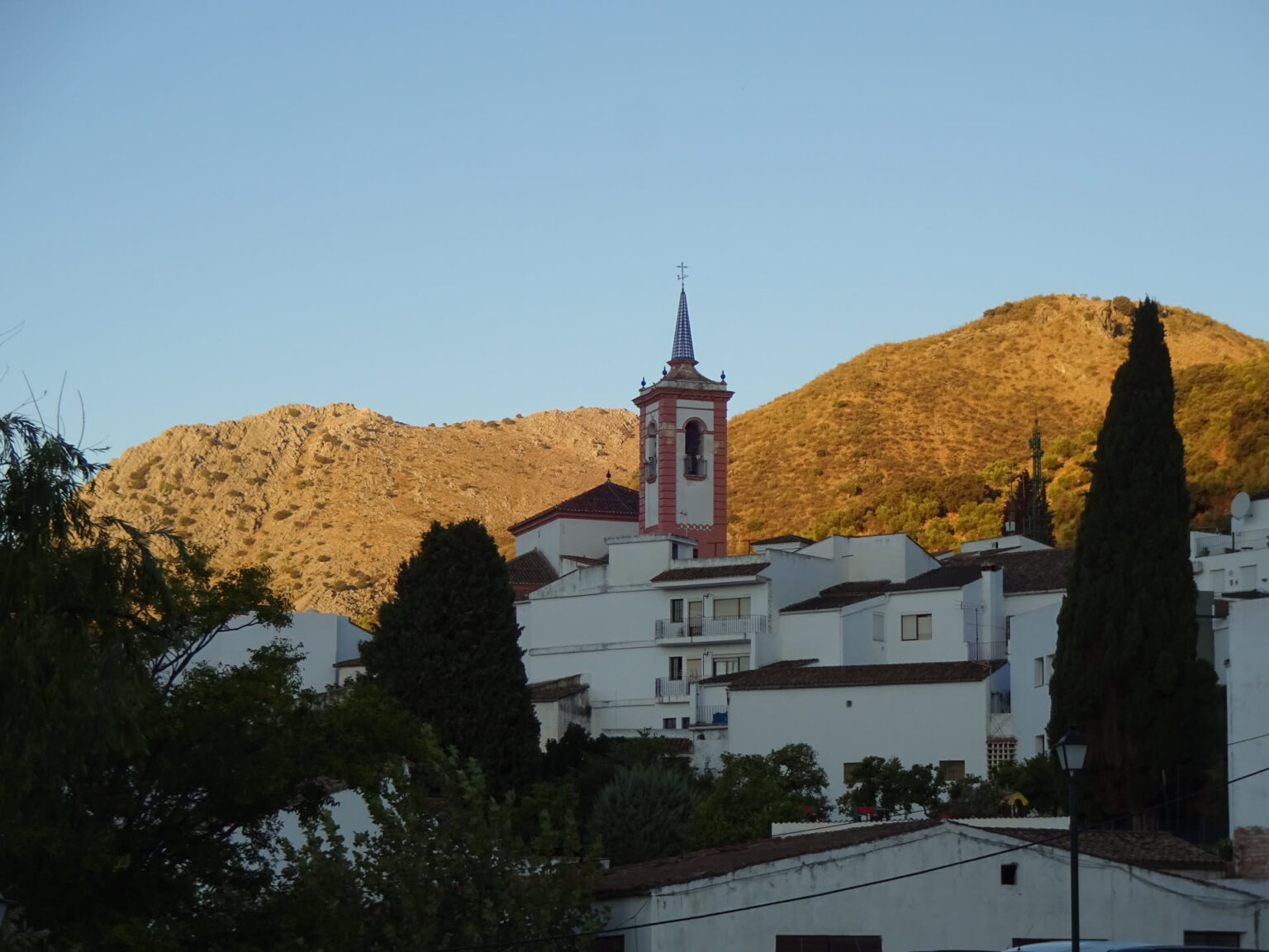 Weißes andalusisches Dorf mit Kirchturm im ersten Morgenlicht, dahinter die golden beleuchteten Hänge