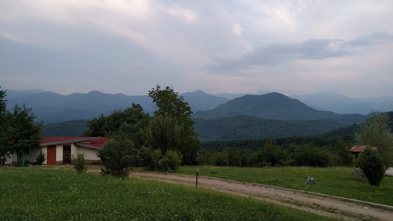 Morgenstimmung vor dem Start: Wiese und Schotterweg vor einem Haus, dahinter die Kette des Balkangebirges unter tief hängenden Wolken