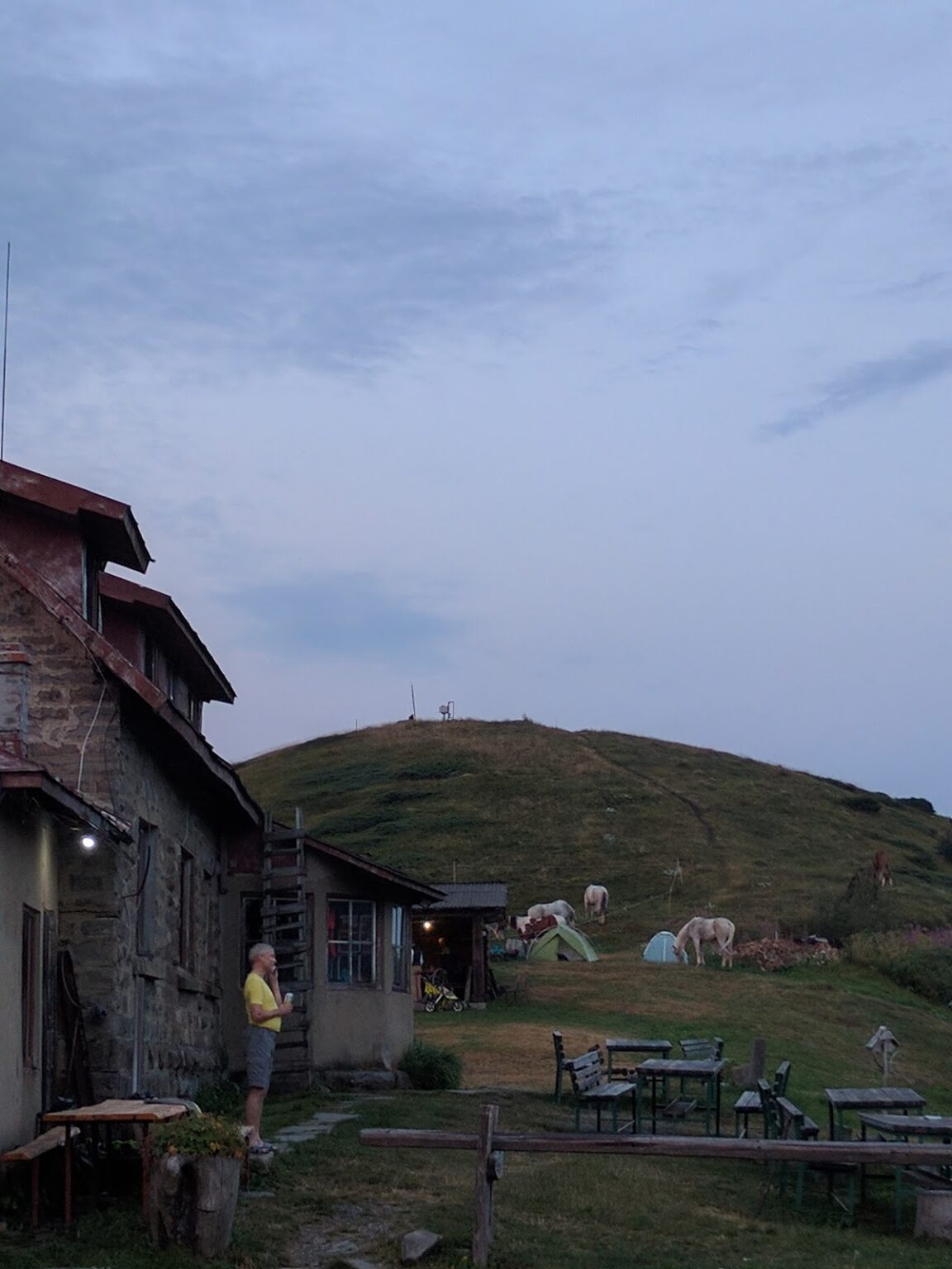 Die Berghütte in der Abenddämmerung, auf der Wiese davor grasen die Pferde neben Bänken und Tischen