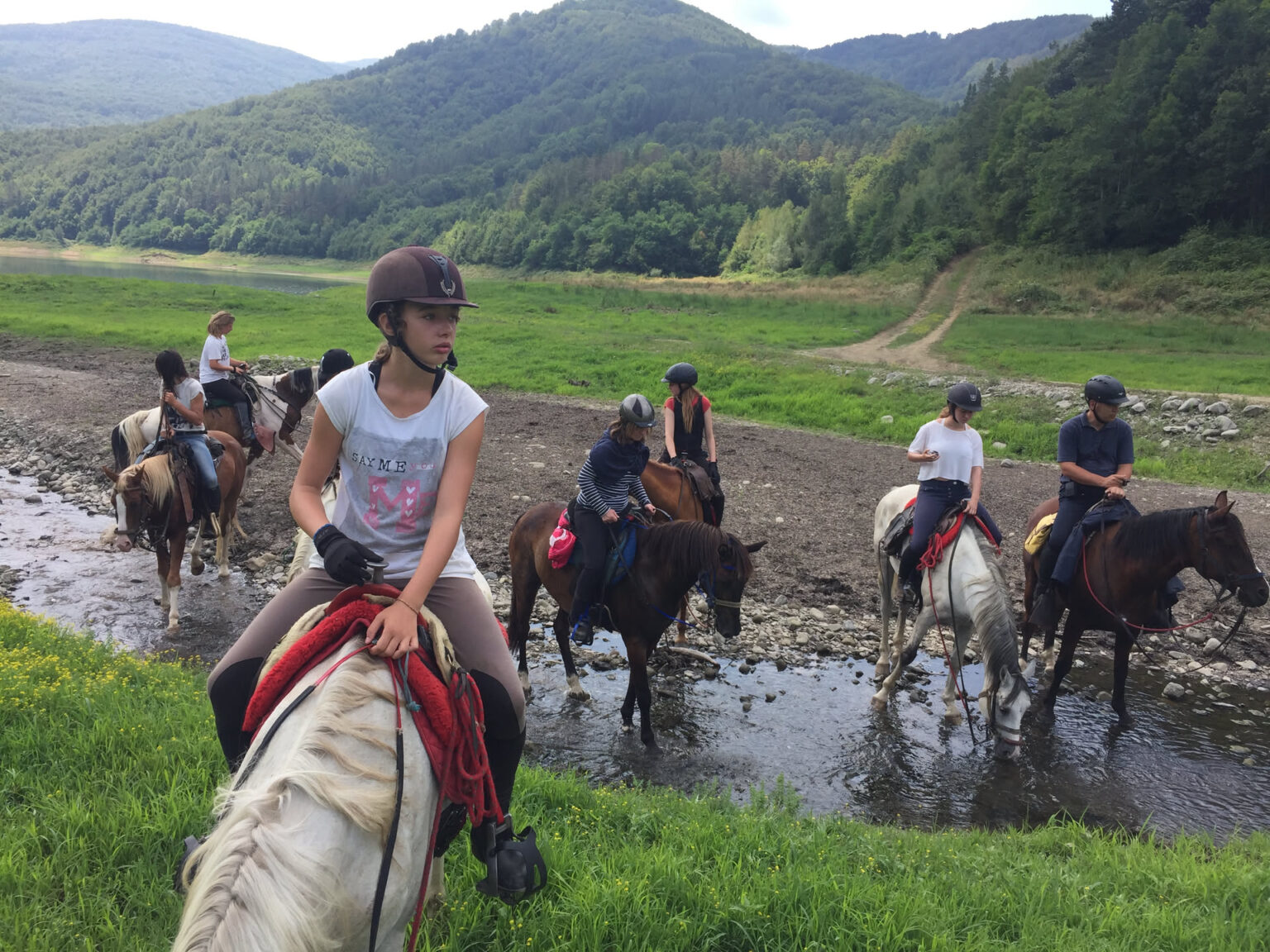 Die Reitgruppe quert ein flaches, breites Flussbett, die Pferde stehen bis über die Fesseln im Wasser