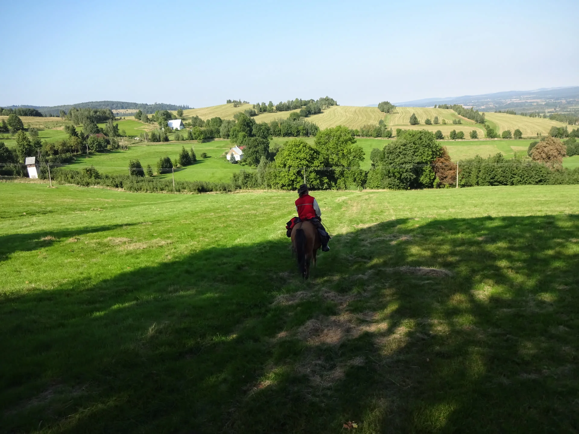 Reiter in roter Weste auf einem braunen Pferd auf einer abschüssigen Wiese, dahinter die hügelige Landschaft des Isergebirges mit Feldern, Baumgruppen und einzelnen Häusern