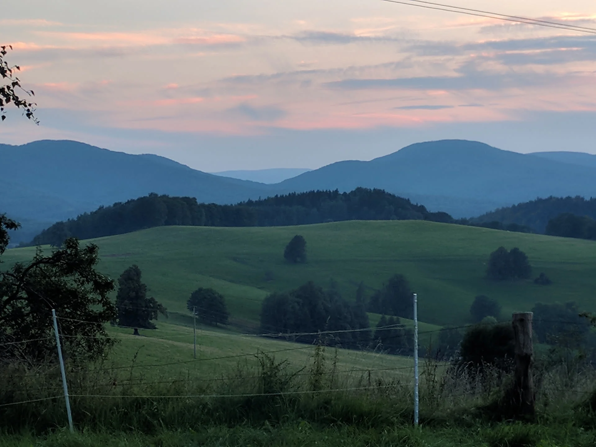 Abendstimmung über grünen Hügeln mit einzelnen Bäumen, dahinter gestaffelte Bergketten unter rosa gefärbtem Himmel, im Vordergrund ein Weidezaun